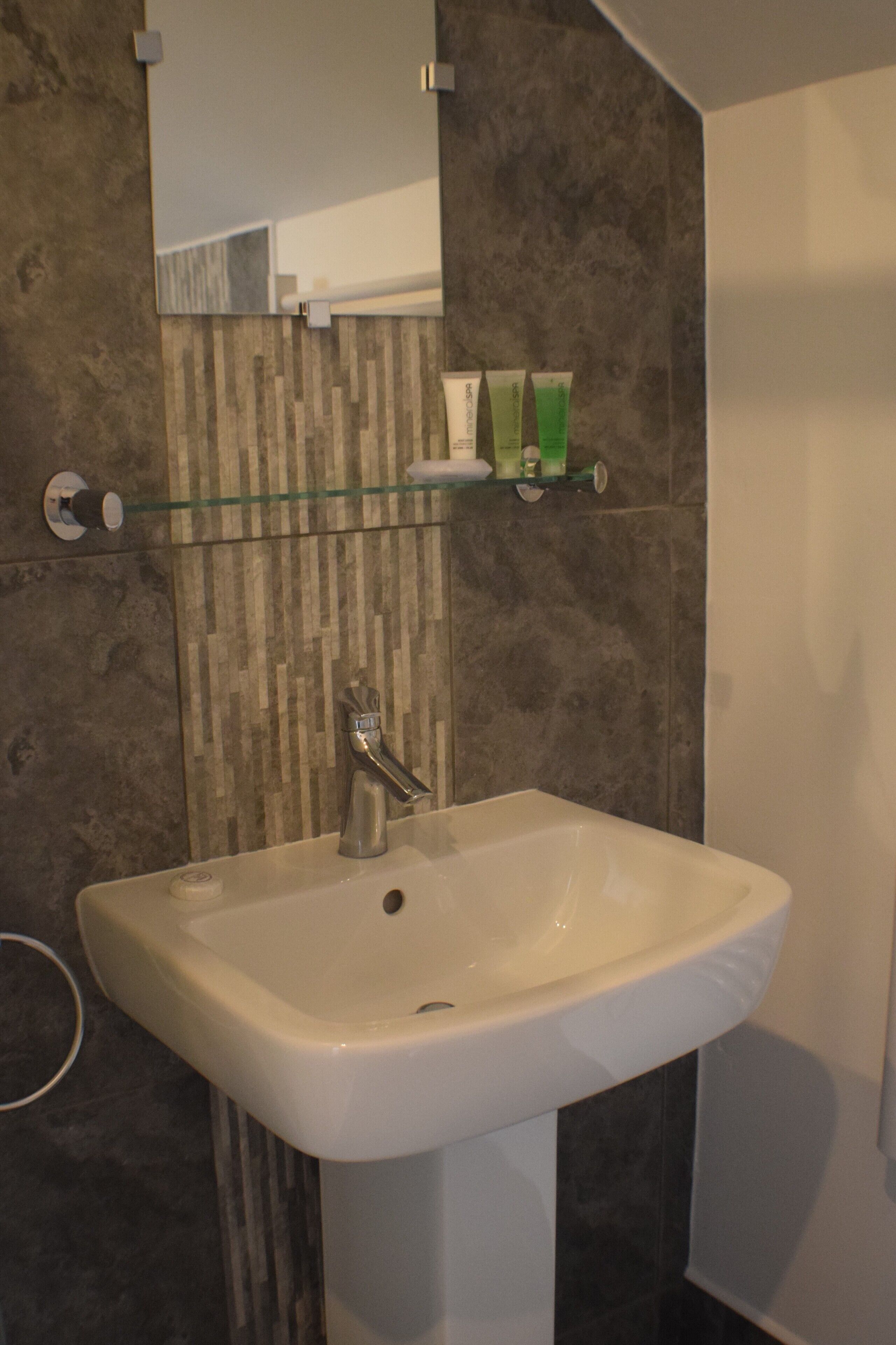 Et badeværelse med en hvid vask, et spejl og en glashylde med toiletartikler.