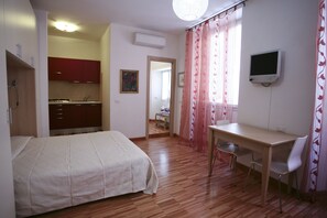 A bedroom with a bed, a desk, and a TV. There is a kitchen area visible through an open door.