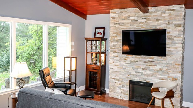 A living room with a stone fireplace, a flat-screen TV mounted on the wall, and a large window offering a view of greenery. - 4-room house with plenty of character (Silverwood)