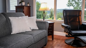 A living room with a grey sofa, a black leather chair, a lamp, and a view of trees through large windows. - 4-room house with plenty of character (Silverwood)