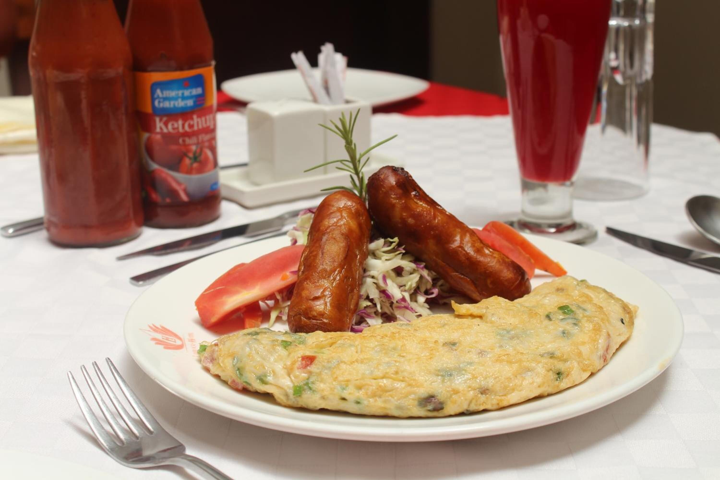 A plate with sausages, coleslaw, and an omelette, with ketchup and a glass of red drink on the table.