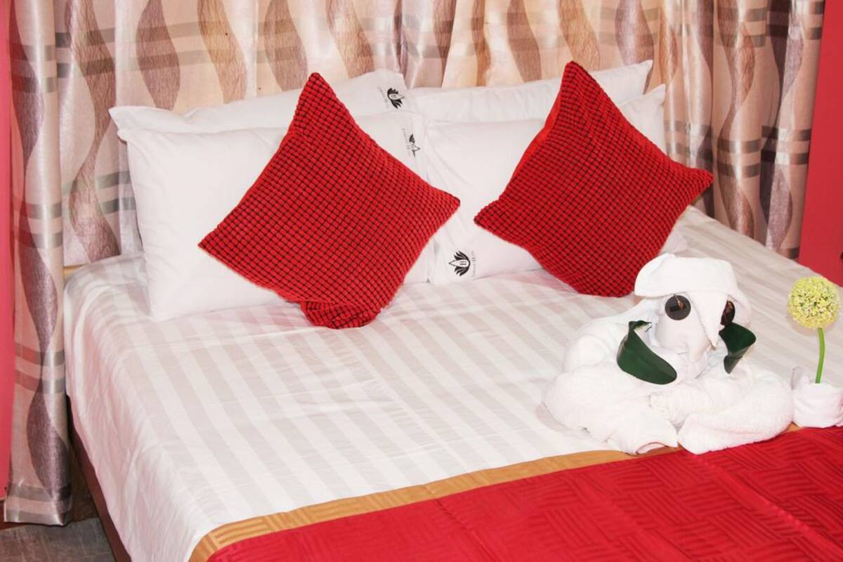 Una cama bien hecha con ropa blanca y dos almohadas rojas, un peluche blanco y una sola flor.