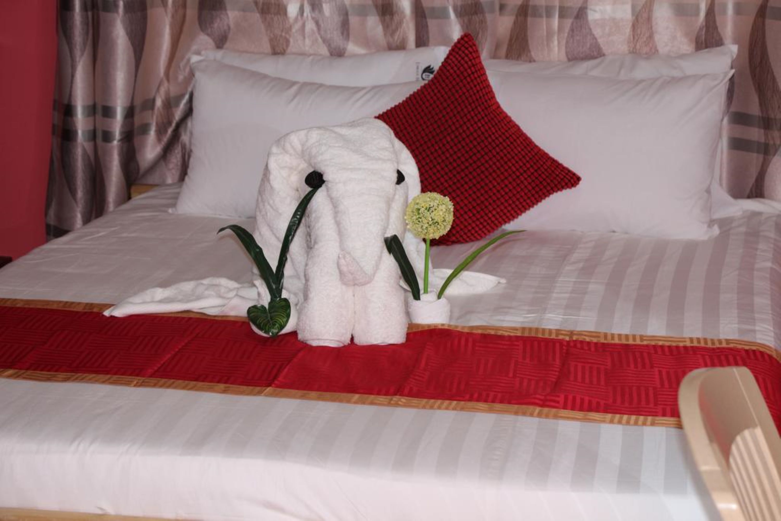 A neatly made bed with a red and white checkered blanket, a red pillow, and a white stuffed animal resembling an elephant. There are two potted plants on the bed.