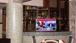 A bar area with a television displaying a news program, shelves of liquor bottles, and a marble column. - Limrock Hotel (Machakos)