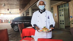 A chef in a white uniform holding a plate with food, standing in front of a red table with a bottle and a glass. - Limrock Hotel (Machakos)