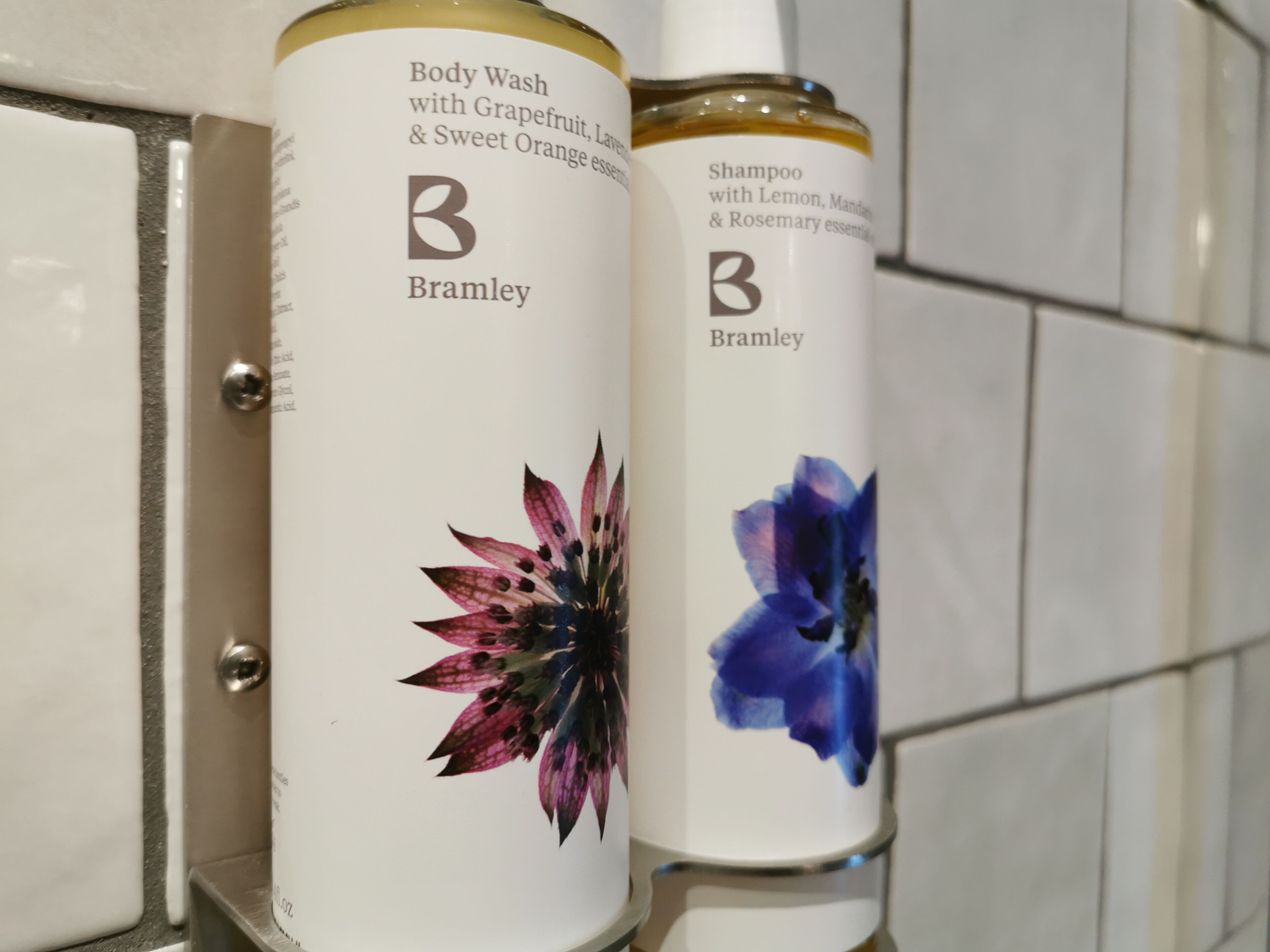 Twee flessen Bramley body wash en shampoo met bloemmotieven, geplaatst tegen een betegelde muur.