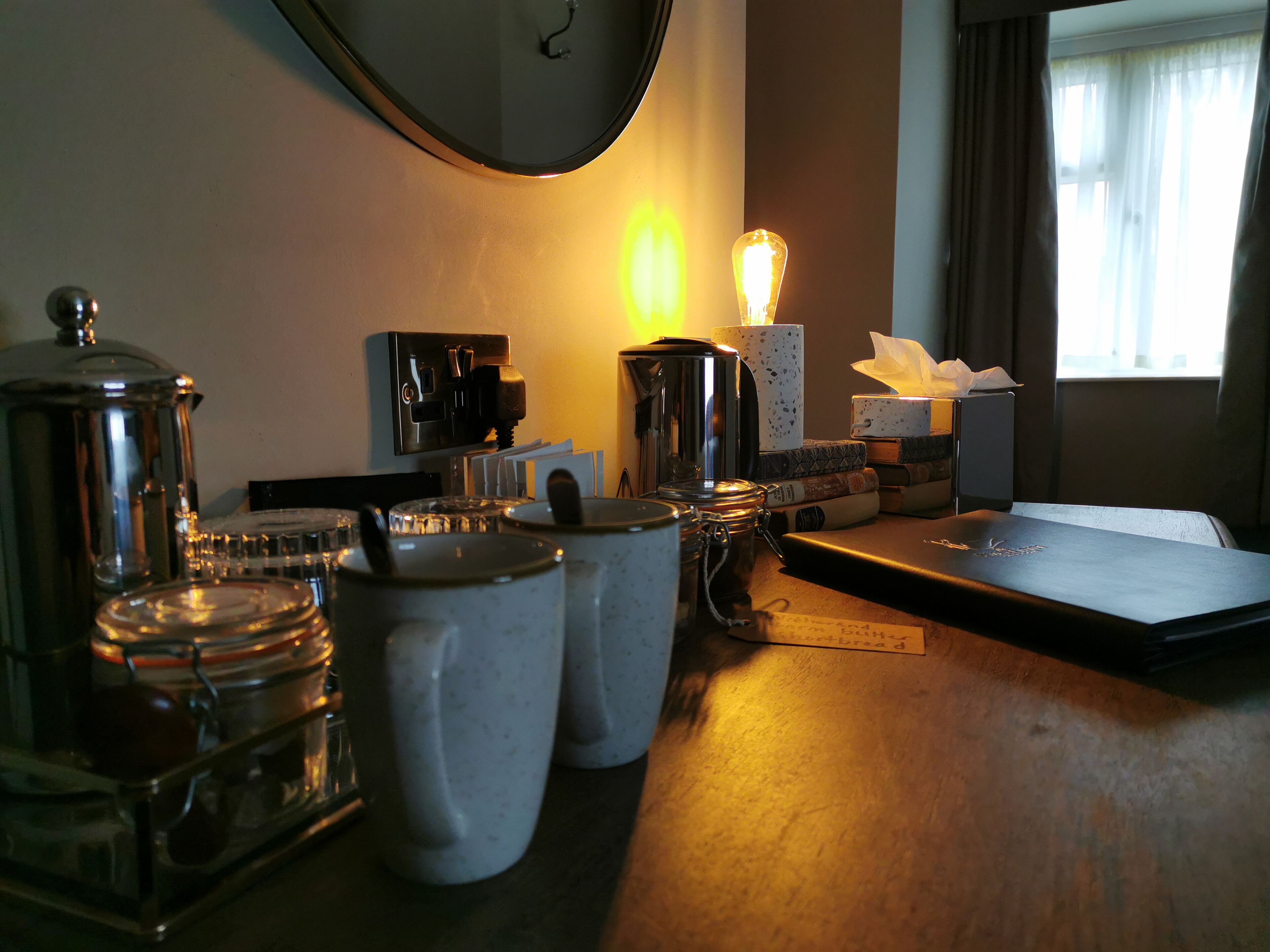 A hotel room with a wooden table, a coffee machine, a lamp, and a window with curtains.