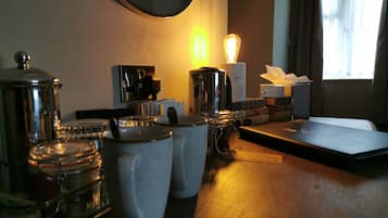 A hotel room with a wooden table, a coffee machine, a lamp, and a window with curtains.