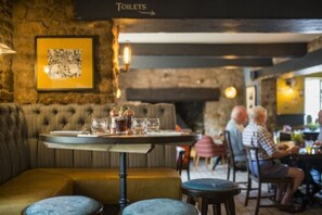 A restaurant with a stone wall, a framed artwork, a table with glasses and a menu, and a sign indicating the location of toilets. - The Swan Inn (Winscombe)