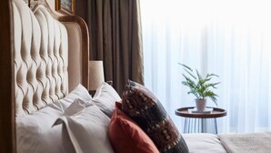 Ein ordentlich bezogenes Bett mit einer gebogenen Kopfteil, ein kleiner runder Tisch mit einem Blumentopf und Blick ins Freie durch transparente Vorhänge.