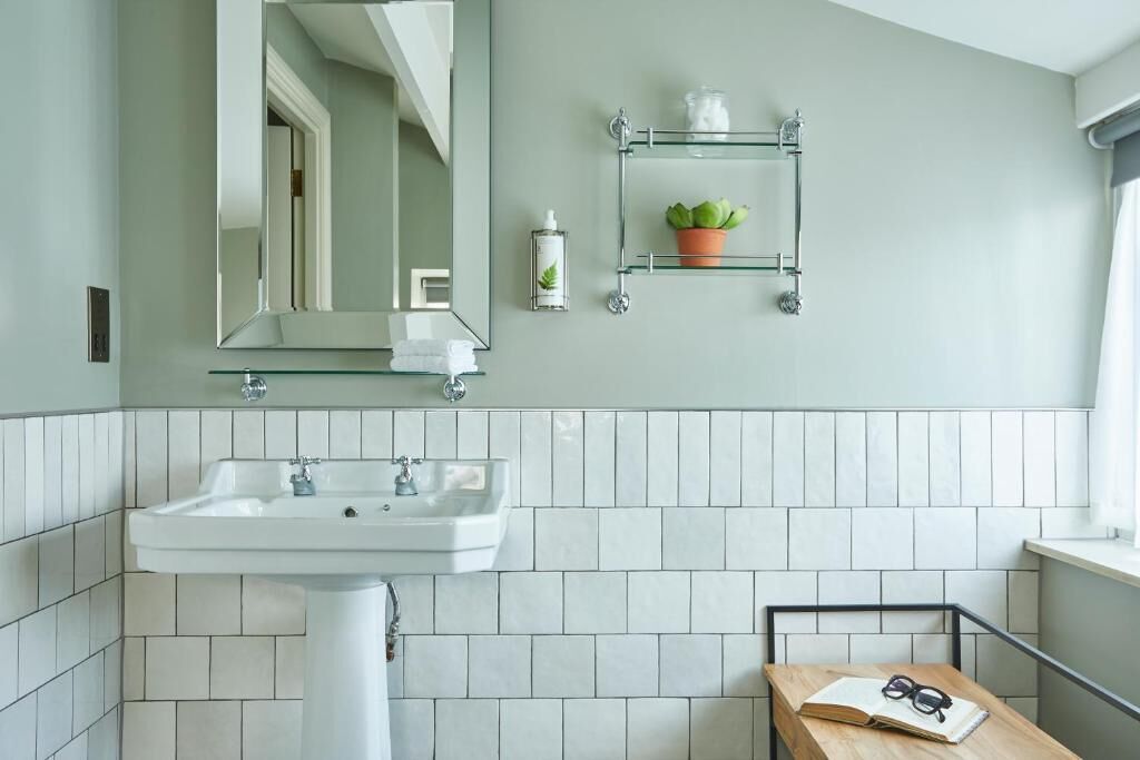 Een badkamer met een wit wastafelmeubel, een spiegel, een glazen plankje met toiletartikelen en een potplant.
