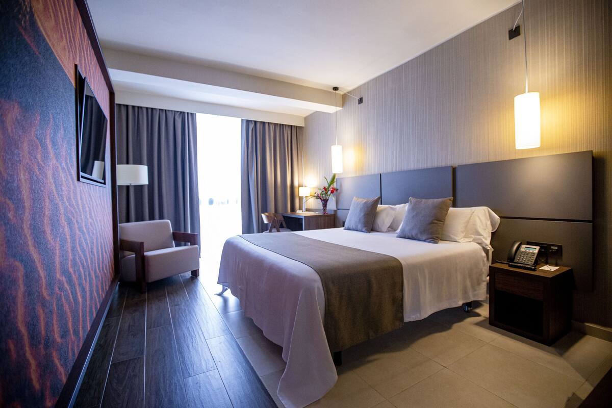 A modern hotel room with a large bed, a desk, a chair, and a nightstand.