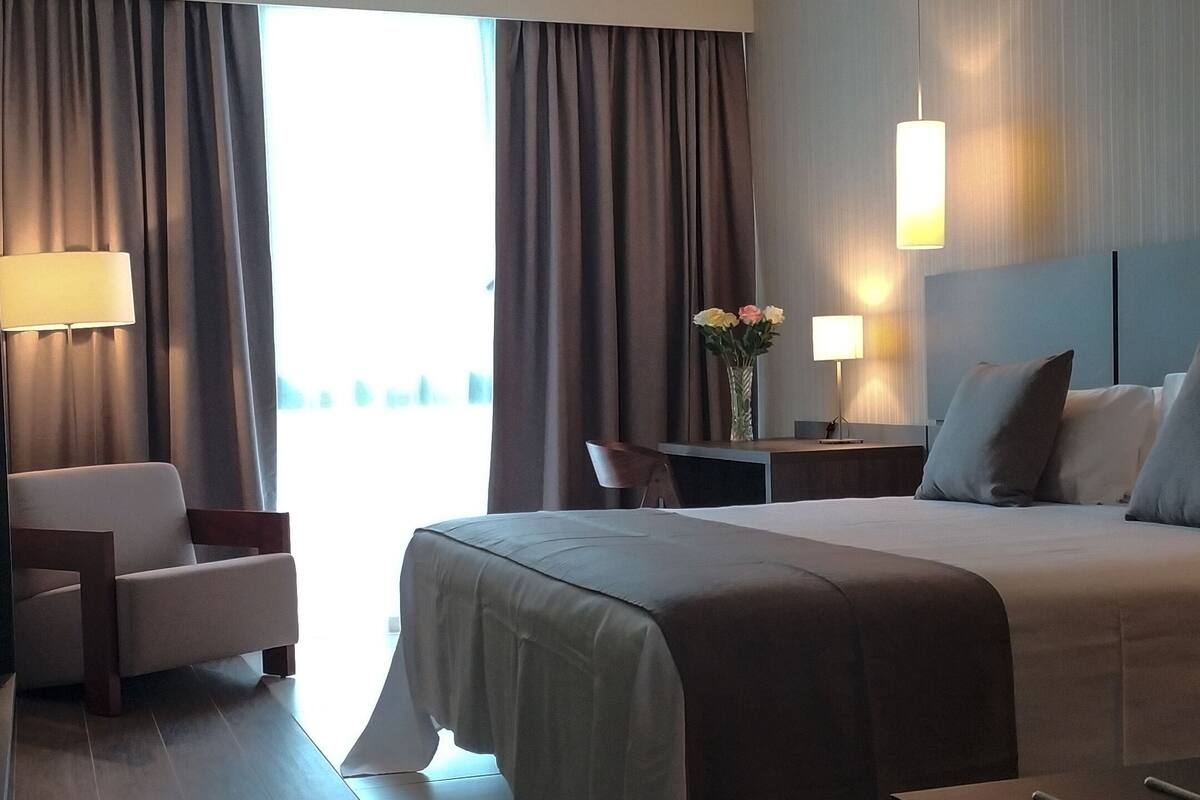 A modern hotel room with a large bed, a desk, a chair, and a view of the city.