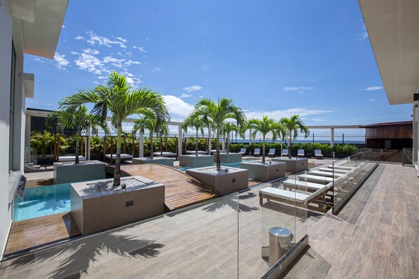 A rooftop pool area with multiple lounge chairs and palm trees, offering a view of the ocean.