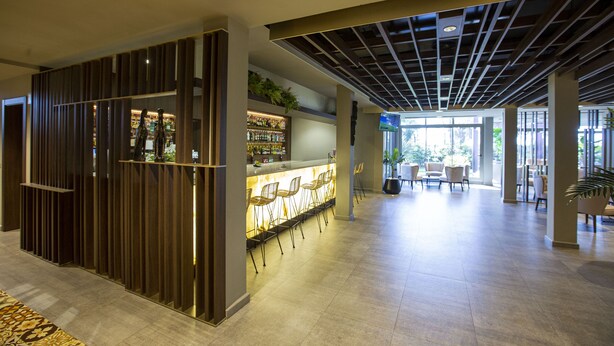 A modern bar area with a long counter and multiple bar stools, a high ceiling with a grid pattern, and a spacious seating area with tables and chairs. - Panafrica Hotel Boutique & Spa (Bata)