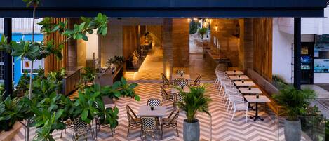 An outdoor dining area with multiple tables and chairs, surrounded by potted plants and a chevron-patterned floor.