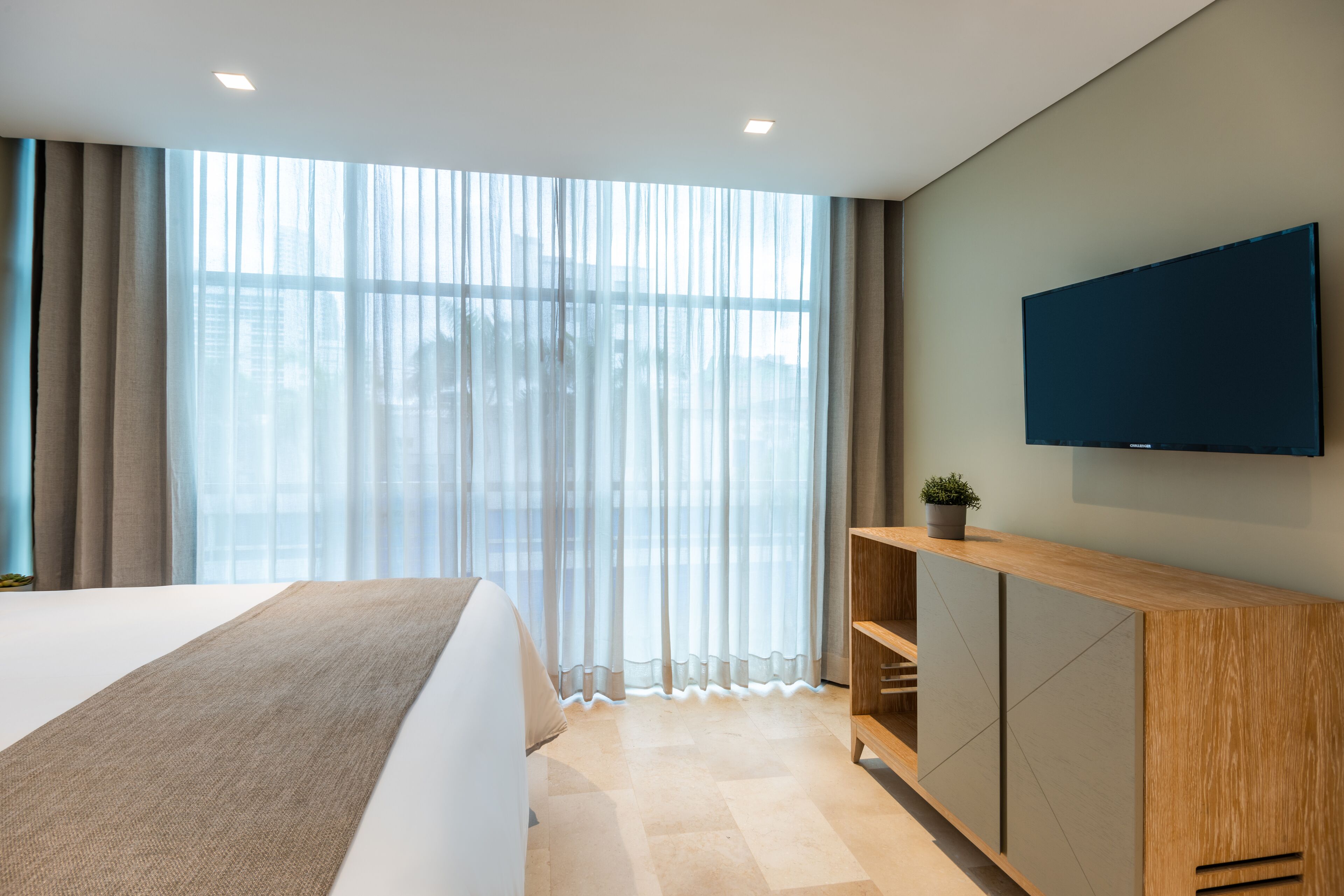 Una habitación de hotel moderna con una cama grande, una mesita consola de madera, un televisor de pantalla plana y un amplio ventanal con cortinas traslúcidas.