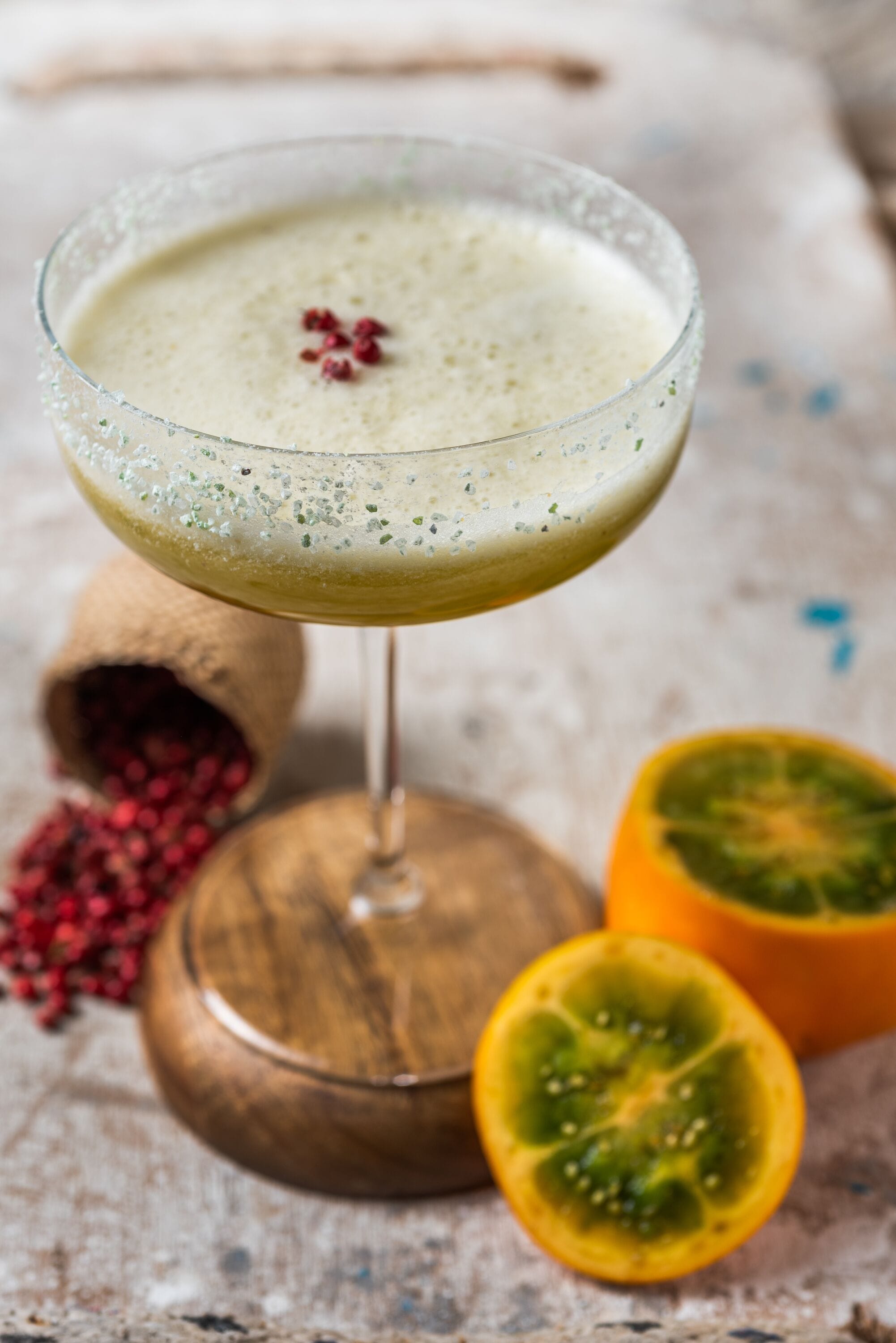 Ein Cocktail im Glas mit Holzunterlage, umgeben von einer halben Orange und einem Jutbeutel mit roten Samen.