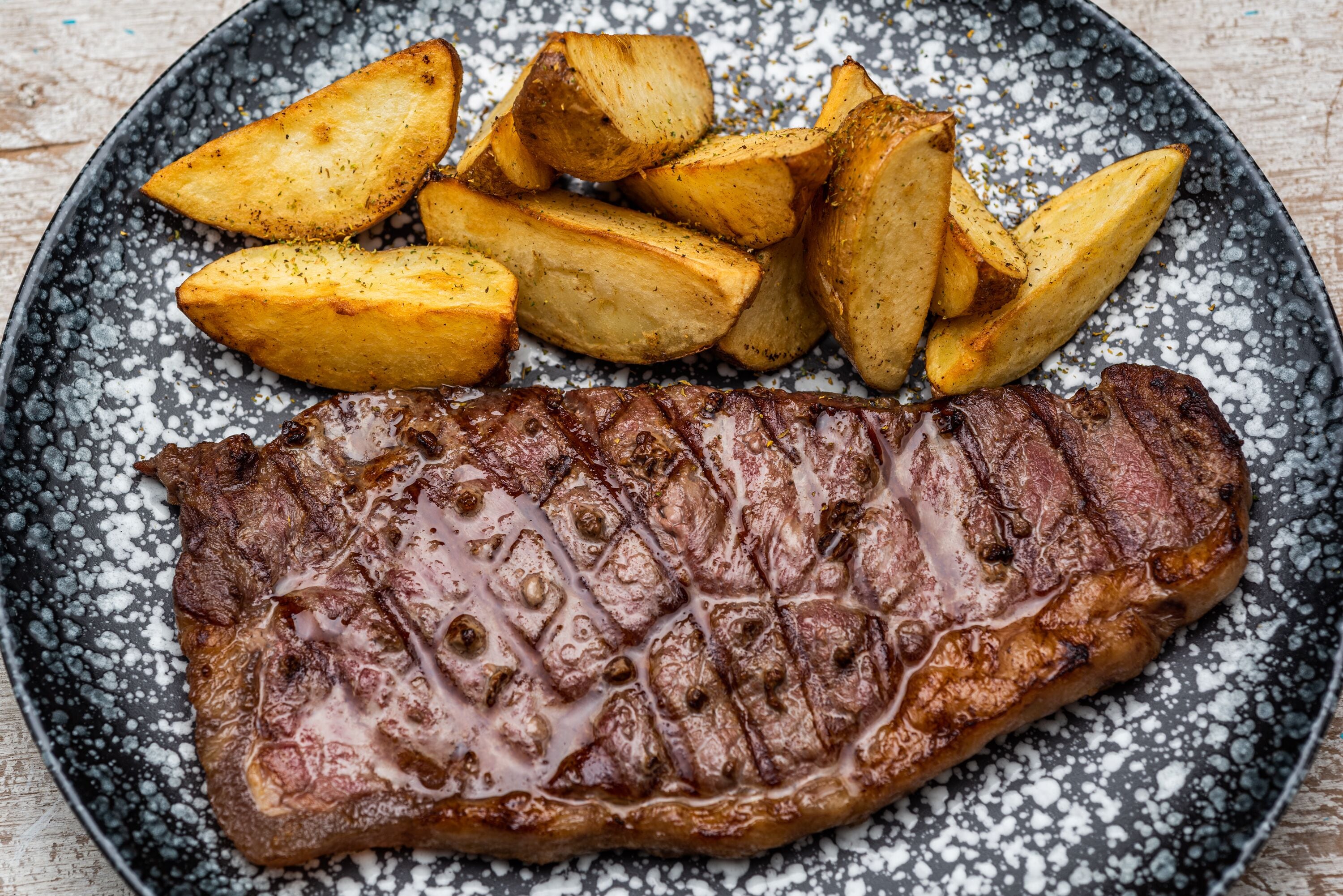 A steak with a crusty exterior and seasoned potatoes on a speckled plate.