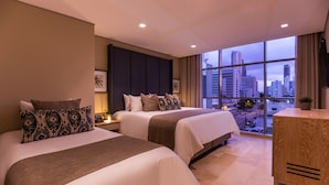 A hotel room with two beds, a large window with a city view, and a small wooden side table.