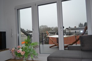 Ein Wohnzimmer mit einer Couch, einem Fernseher und Blick auf einen Balkon mit einer Hängematte und einem Zaun.