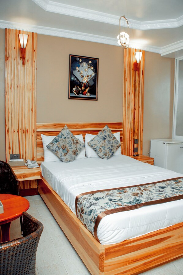 A hotel room with a wooden bed, a small table, a chair, and a painting on the wall.