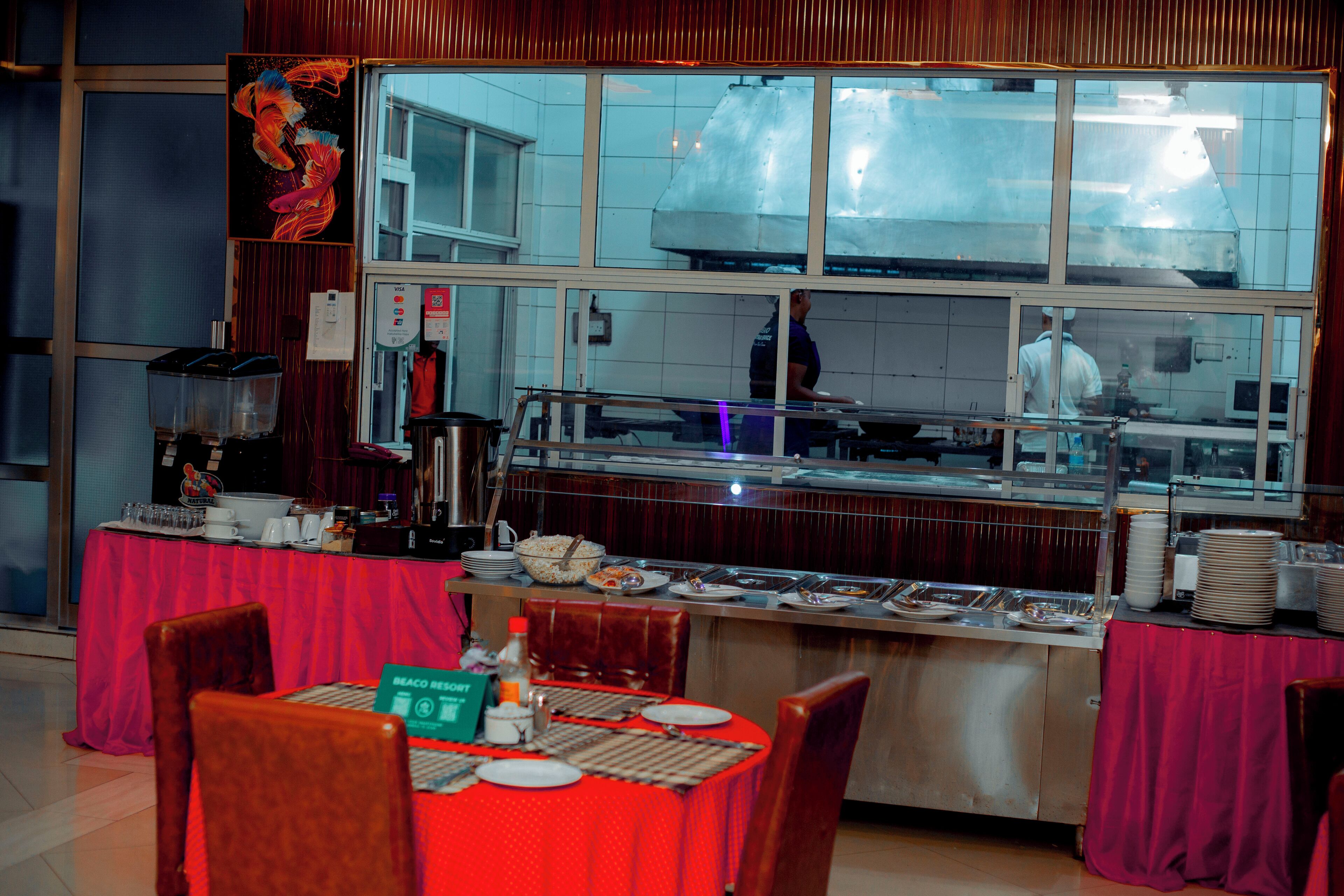 A buffet setup with a stainless steel counter, red tablecloths, and a large window overlooking the kitchen.