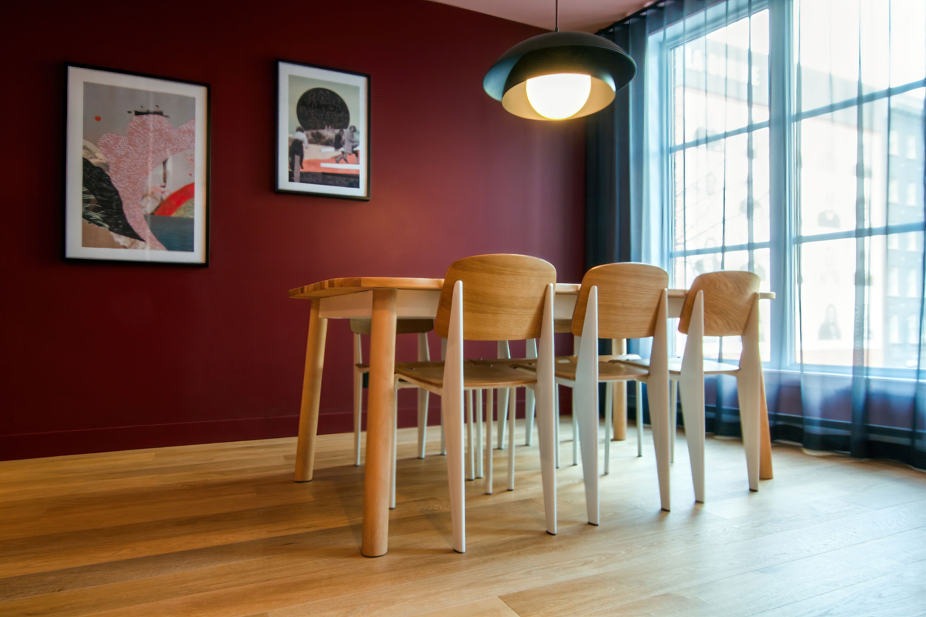 A dining area with a wooden table and chairs, framed artwork on the wall, and a large window with natural light.