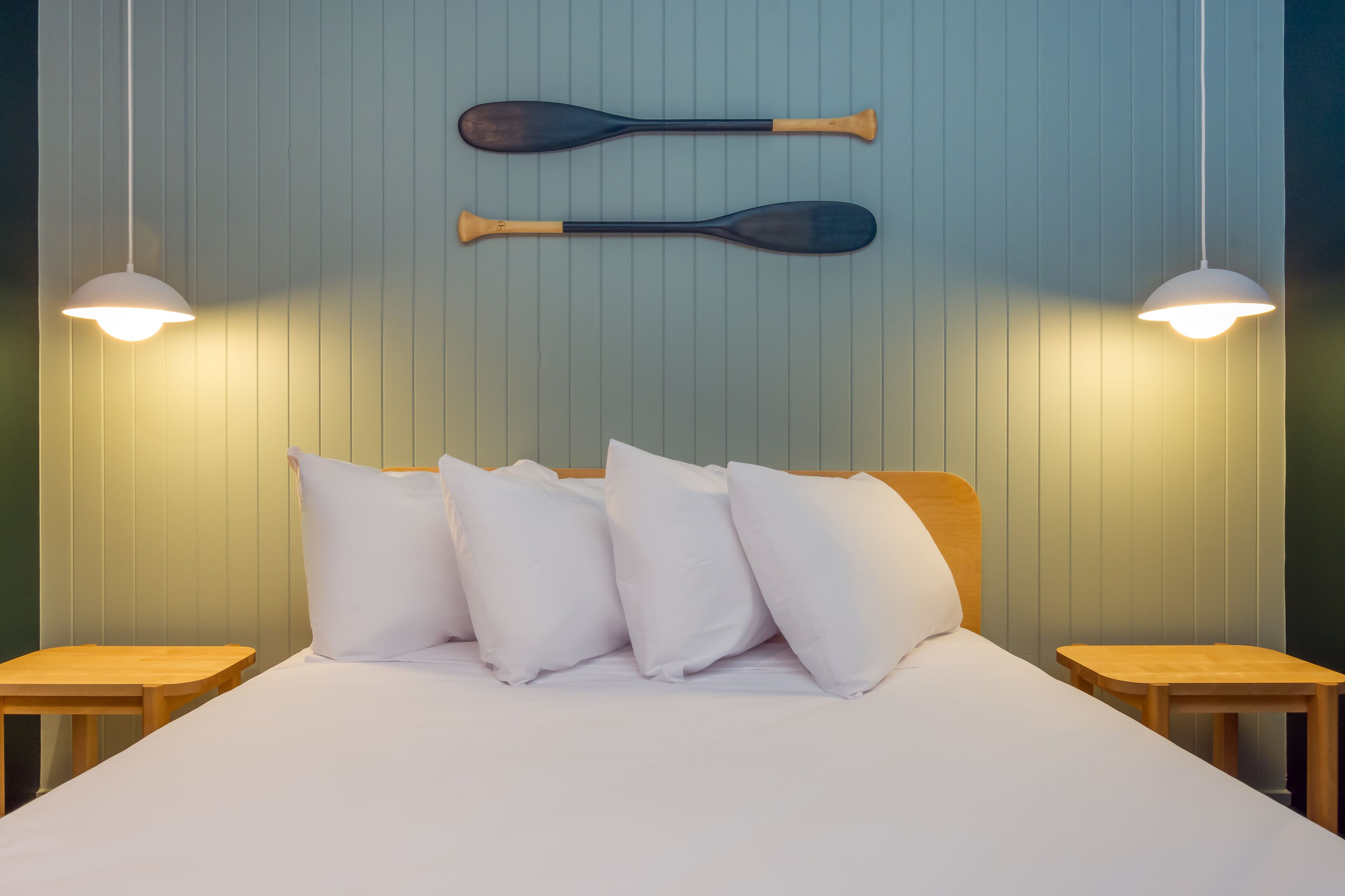 A neatly made bed with white bedding and several pillows, two wooden nightstands on either side, and two paddles hanging on the wall above the bed.