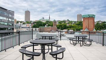 A rooftop terrace with multiple tables and chairs, overlooking a cityscape with buildings and a church spire.