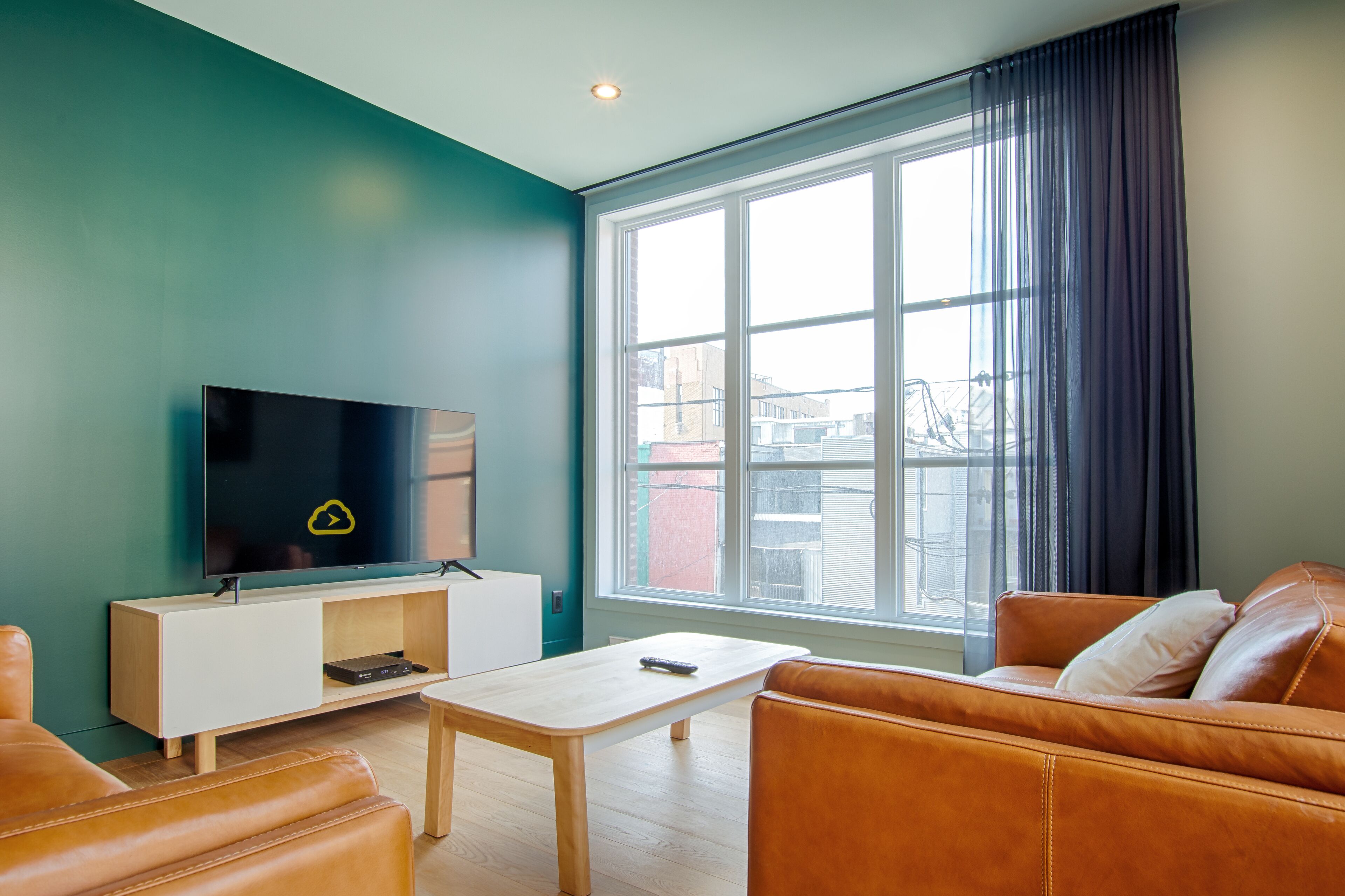 Una sala de estar moderna con un ventanal grande, un televisor de pantalla plana, un mueble blanco para entretenimiento y un sofá de cuero marrón.