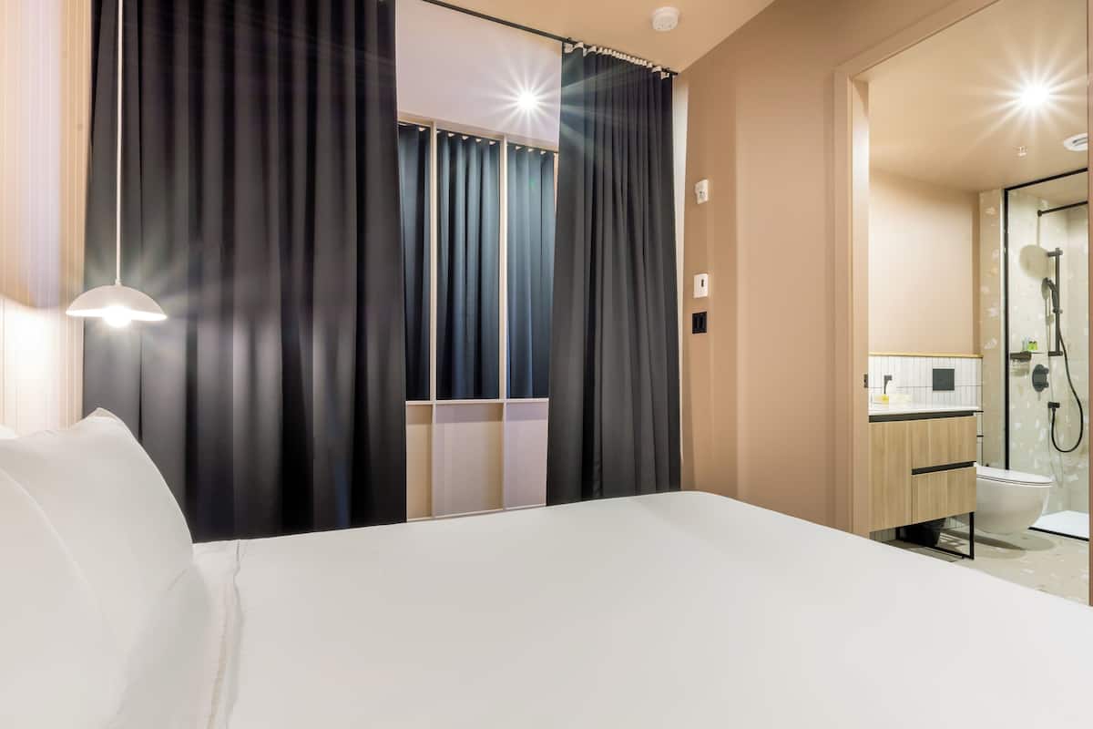 A hotel room with a bed, a nightstand, a mirror, a shower area, and a window with curtains.