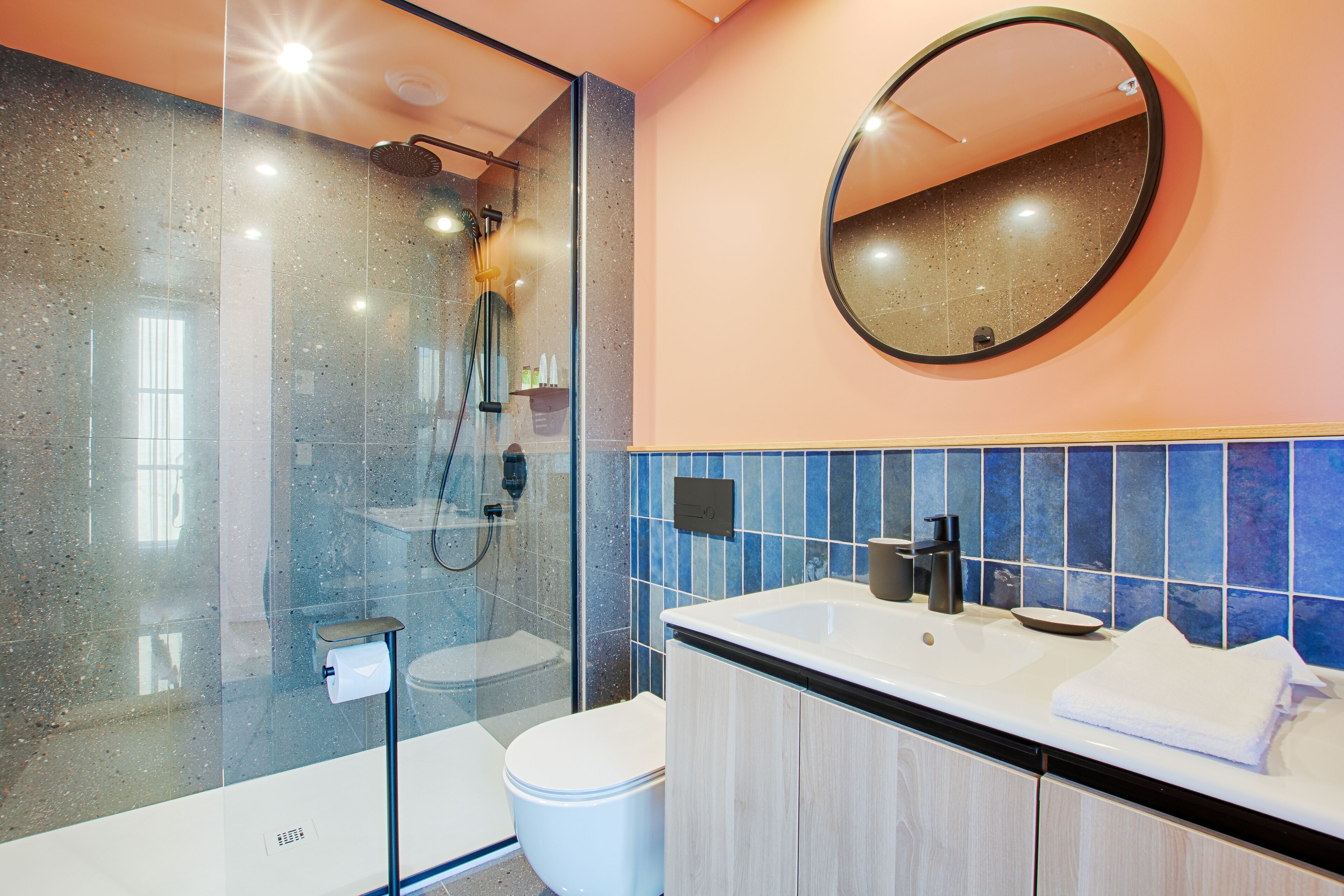 Un baño moderno con ducha envuelta en vidrio, inodoro blanco, lavamanos con espejo y paredes de azulejos azules.