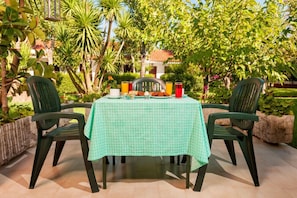 An outdoor dining area with a table set for two, surrounded by greenery and a building in the background. - Alykes Park Bungalows & Apt (Zakynthos)
