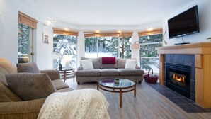 A cozy living room with a fireplace, a sofa, a coffee table, and large windows offering a view of trees.