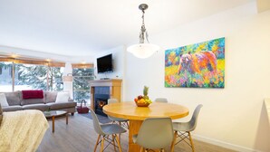 A modern living room with a sofa, a dining table with chairs, a fireplace, and a large window with a view of trees.