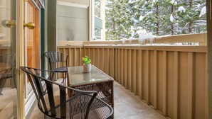A balcony with a glass table and chairs, a potted plant, and a view of snow-covered trees.