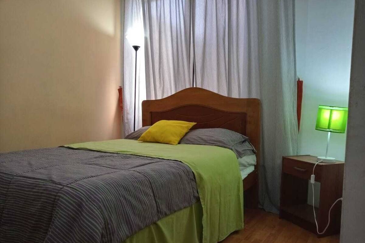 A bed with a wooden headboard, a green and gray striped bedspread, and a yellow pillow. There is a nightstand with a green lamp beside the bed.
