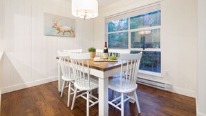 A dining area with a wooden table, white chairs, a chandelier, a painting of an elk, and a window with a view of trees. - Luxury 2BR/2BA – Next to slopes, Private Garage (Whistler)