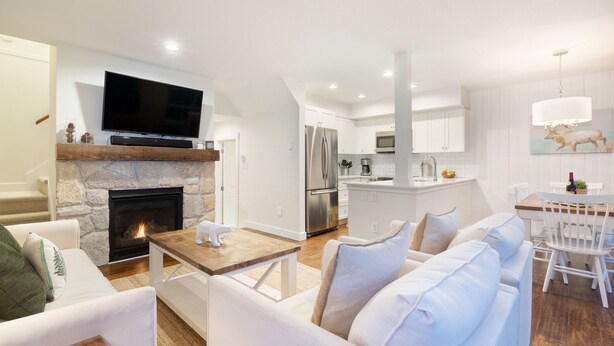 A modern living room with a fireplace, a flat-screen TV, and a dining area with white furniture. - Luxury 2BR/2BA – Next to slopes, Private Garage (Whistler)