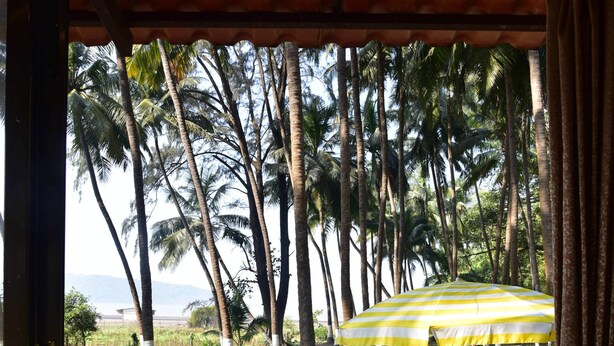 An outdoor patio with stone flooring, a yellow and white striped umbrella, and palm trees in the background. - Beach Cabana - La Mer Beach House (Revdanda)