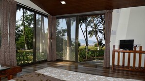 A room with a large sliding glass door leading to a balcony with a view of palm trees and a beach. There is a bed with a floral bedspread and a wooden bench. - Beach Cabana - La Mer Beach House (Revdanda)
