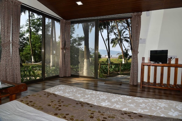 A room with a large sliding glass door leading to a balcony with a view of palm trees and a beach. There is a bed with a floral bedspread and a wooden bench.