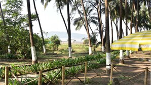 A tropical garden with palm trees, a wooden fence, and a yellow-striped umbrella. - Beach Cabana - La Mer Beach House (Revdanda)