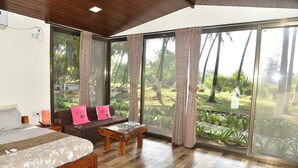 A bedroom with a bed, a sofa, a wooden coffee table, and a large sliding glass door leading to a garden. - Beach Cabana - La Mer Beach House (Revdanda)