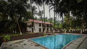 A rectangular pool with a stone deck, surrounded by palm trees and a white building with a red roof. - 104 - Ocean View - La Mer Beach House (Revdanda)
