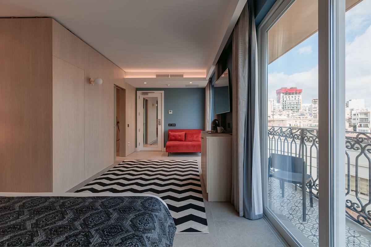 Une chambre d’hôtel moderne avec un grand lit, un canapé rouge, un balcon offrant une vue sur la ville et un tapis à motifs.