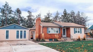 A two-story house with a brick chimney, a small porch, and a front yard with grass and scattered flowers.