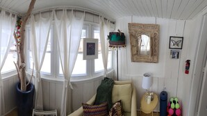 A cozy room with a wooden ceiling, a large window with curtains, a small table with a lamp, and a chair with a colorful cushion.