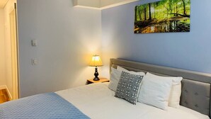 A bedroom with a bed, a nightstand with a lamp, a wall-mounted painting, and a door. - WindTower Resort @ Cozy Hotel Room (Canmore)